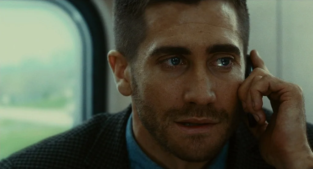 Source Code (2011) movie still: closeup — A passenger on a commuter train sits beside a large rectangular window. A man speaks into a mobile phone…