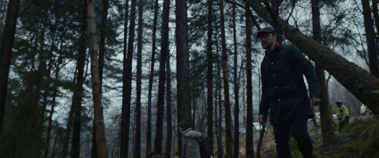 My Son (2021) movie still: medium — A dense woodland area with thin vertical tree trunks and mossy ground texture. A man walks through the forest…