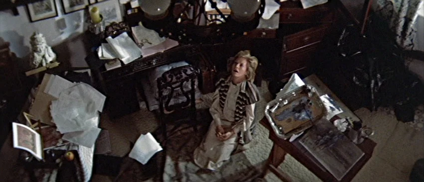 Images (1972) movie still: wide — A cluttered room contains desks covered with loose papers, art supplies, and a small sculpture. A woman kneels on…