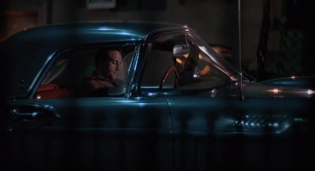 Puzzle of a Downfall Child (1970) movie still: medium — A man sits inside a dark blue 1950s convertible parked on a nighttime street. The vehicle's…