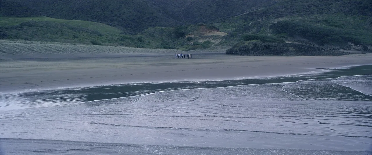 Dirty Little Secret (1998) movie still: wide — A vast, empty beach with dark wet sand meets a calm, light-reflecting ocean tide. A small group of…