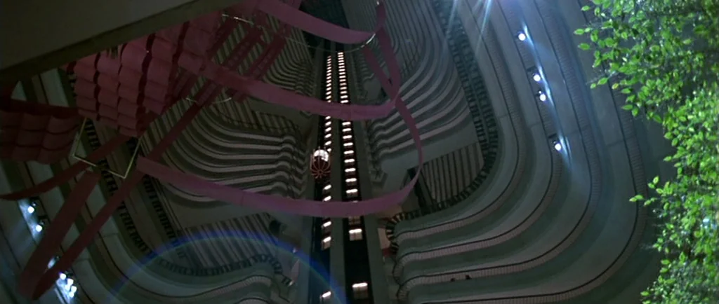 Manhunter (1986) movie still: wide — A massive hotel atrium features curved balconies stretching upward toward a dark ceiling. A single elevator car…