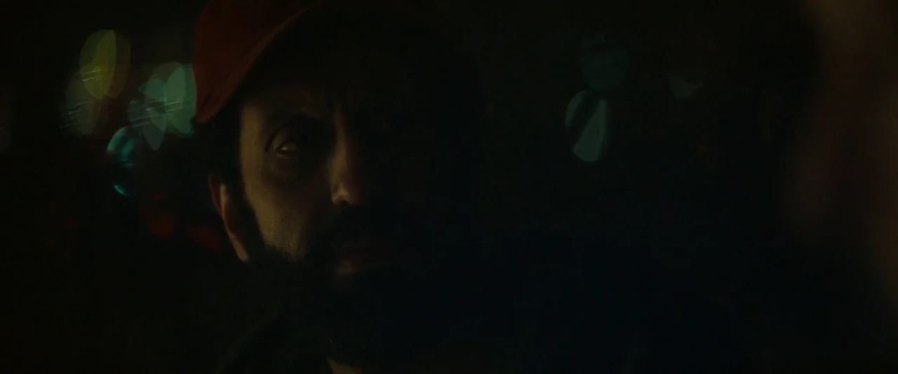 Ali & Ava (2021) movie still: closeup — A man in a red baseball cap stands in a dark city at night. His face is illuminated by faint off-screen…