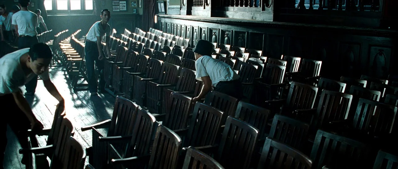 Ip Man 2 (2010) movie still: wide — An interior space contains many rows of wooden chairs arranged on a wooden floor. Young men wearing white…
