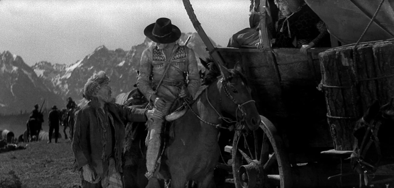 The Big Trail (1930) movie still: wide — A mountain range serves as a backdrop to a wagon train resting in a meadow. An older man stands beside a…