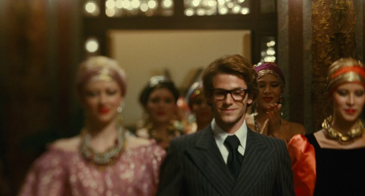 Saint Laurent (2014) movie still: medium — A man in a sharp suit walks through a crowd at an event. Several women in glamorous vintage attire are…