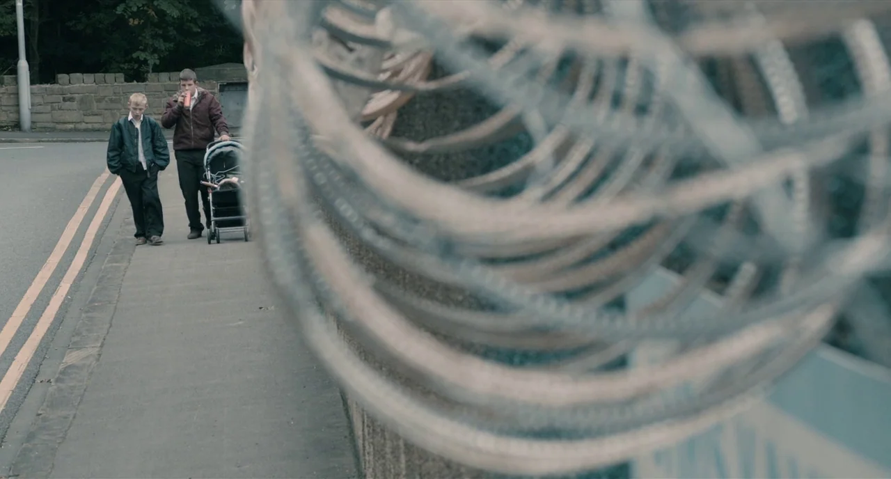 The Selfish Giant (2013) movie still: wide — A paved path runs alongside a stone wall topped with heavy barbed wire coils. Two youths walk together,…