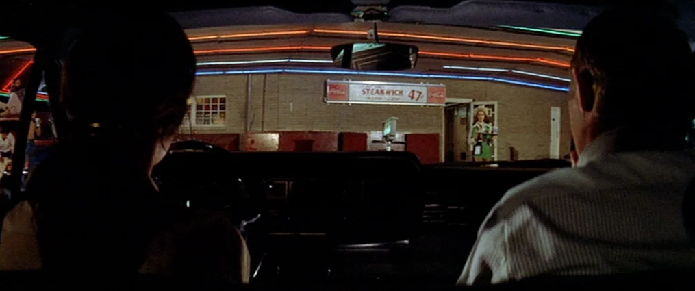 The Getaway (1972) movie still: wide — A night view from inside a car looking toward a 1970s style drive-in restaurant. A carhop server stands in…