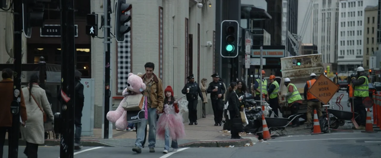 Wolf Man (2025) movie still: fullbody — An urban intersection with construction work and a green pedestrian light overhead. A man holding a pink…