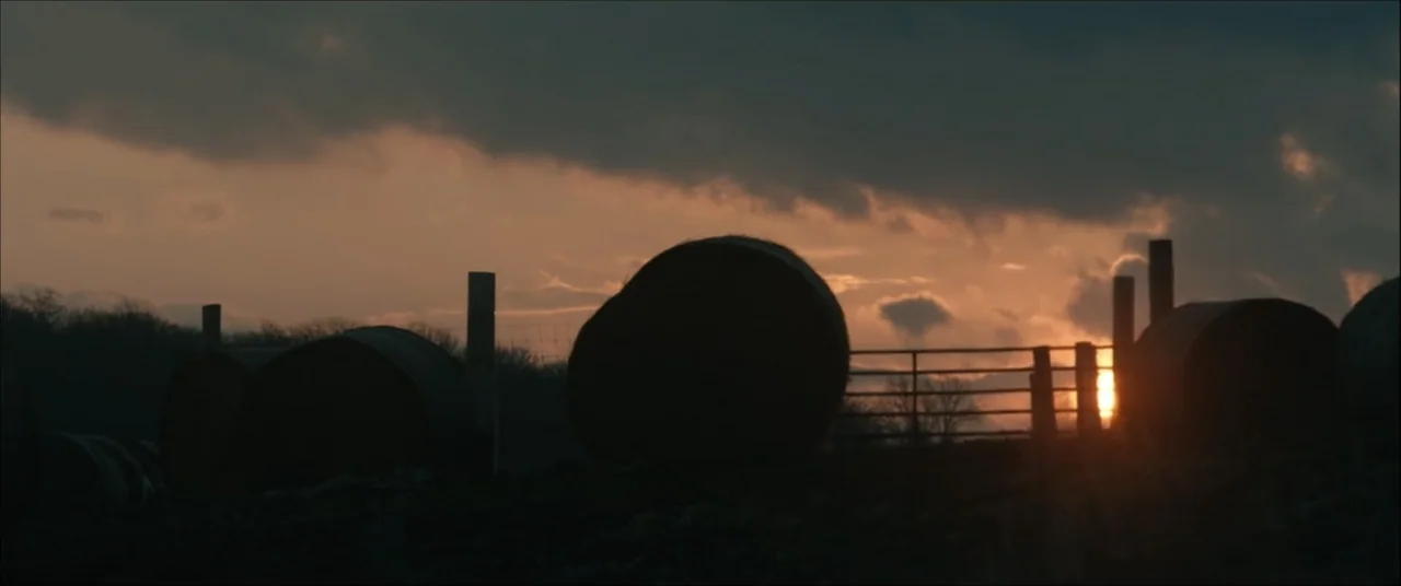 I'll Take Your Dead (2018) movie still: wide — A farm field at sunset with circular hay bales silhouetted against the bright horizon. A metal gate…