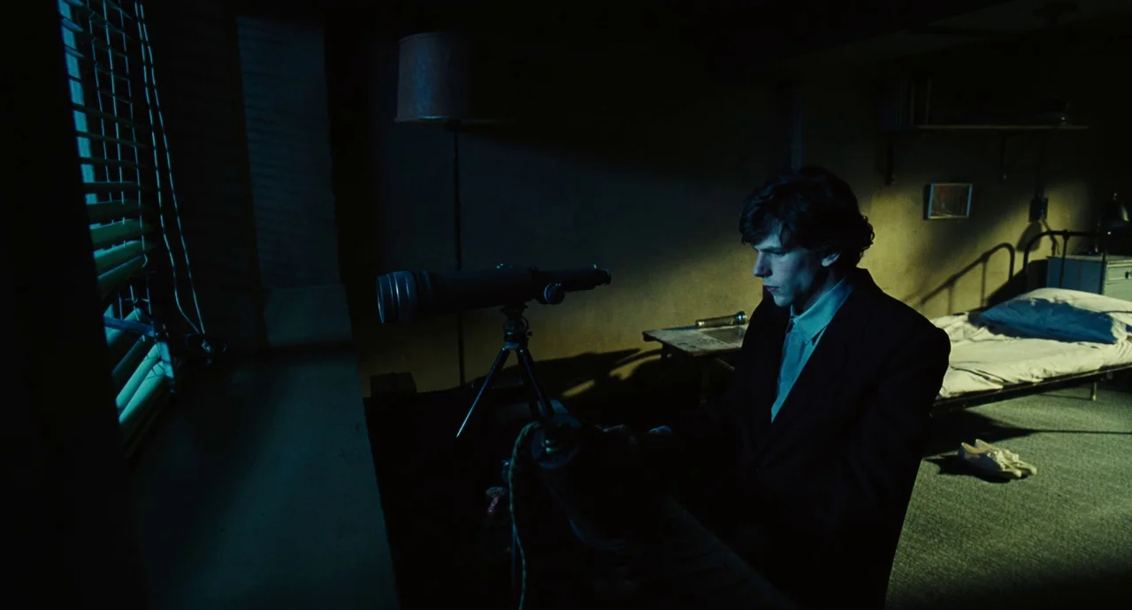 The Double (2013) movie still: medium — A small dark room contains a single metal bed and a cluttered desk. A man wearing a suit sits at the desk…