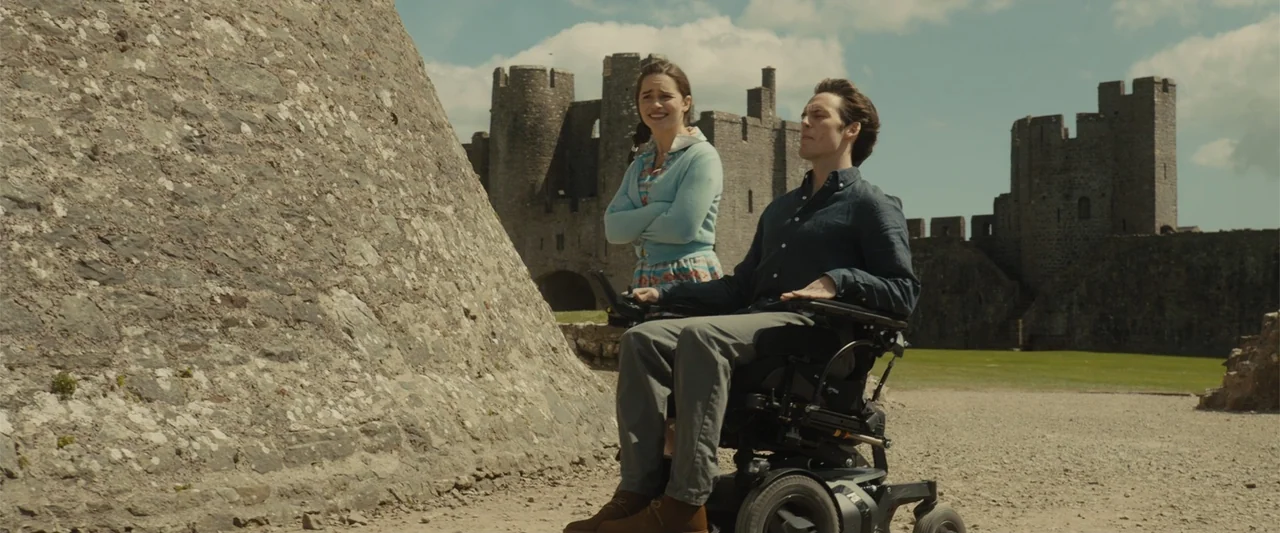 Me Before You (2016) movie still: fullbody — A woman and a man in a wheelchair stand before old stone castle walls. The man remains seated in his…