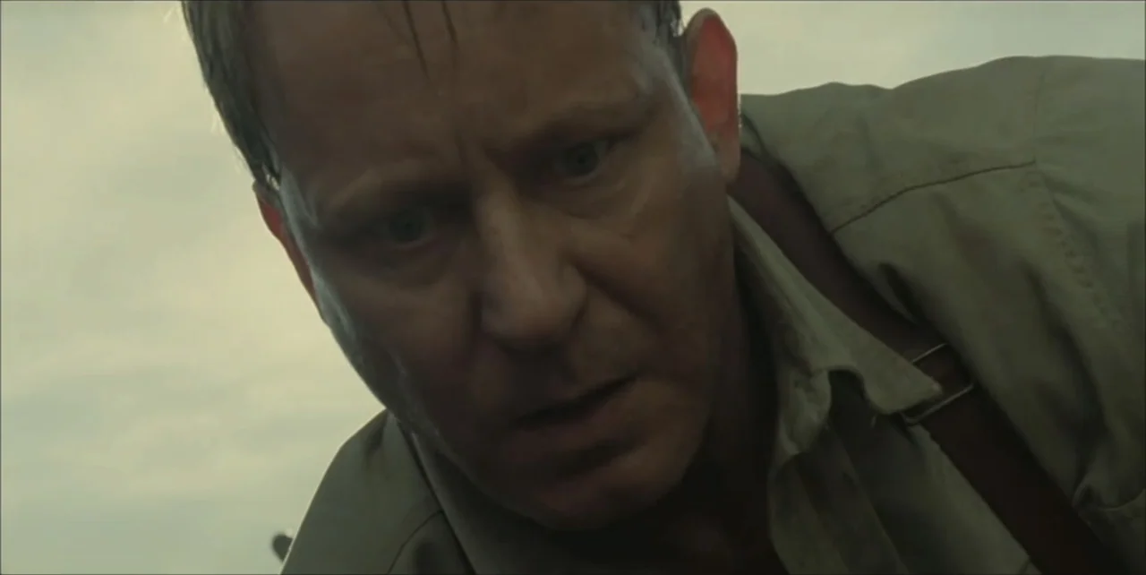 Exorcist: The Beginning (2004) movie still: closeup — An outdoor field provides the background under an overcast, featureless sky. A middle-aged man…