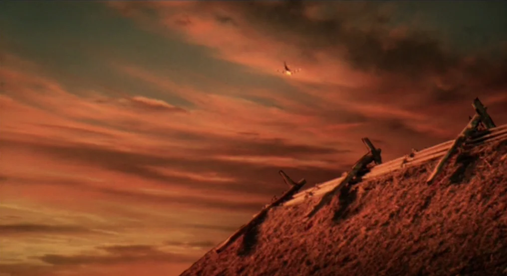 Rhapsody in August (1991) movie still: establishing — A steeply pitched thatched roof is visible against a vibrant, fiery sunset sky. A distant…