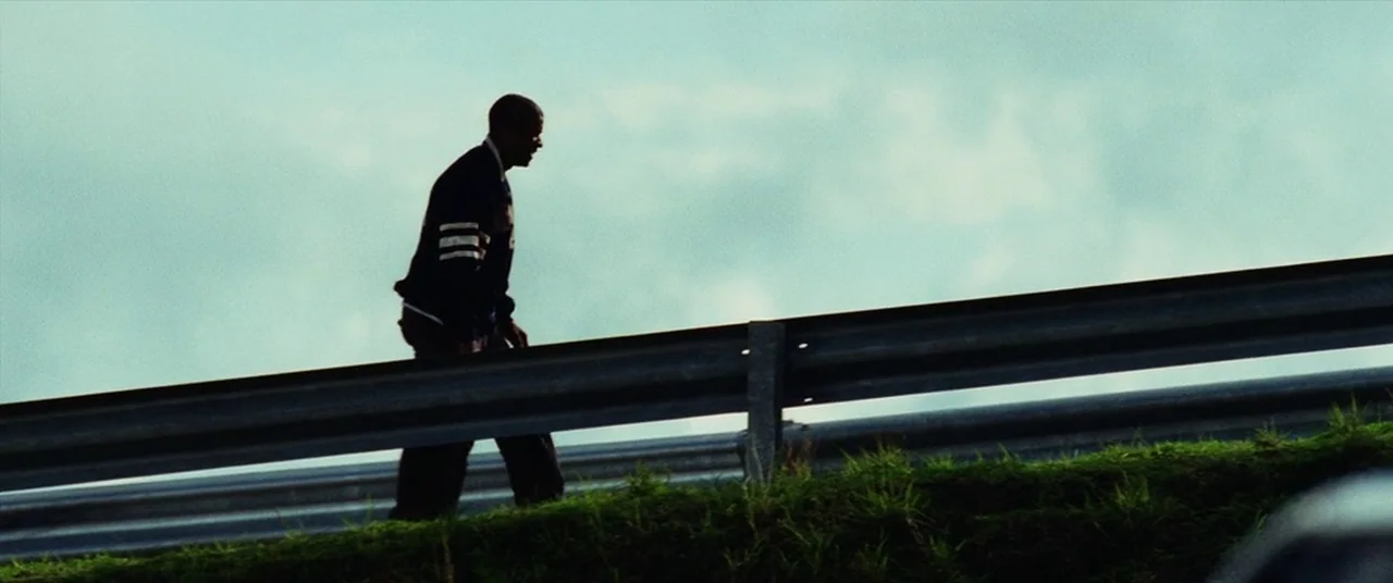 Man on Fire (2004) movie still: fullbody — A man walks along the grassy verge next to a metal highway guardrail. Bright, overcast sky looms behind…