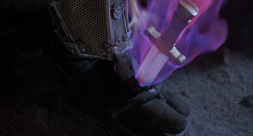 Brazil (1985) movie still: closeup — A dark rocky surface holds a black boot with a metallic mesh accessory. A glowing purple dagger stands upright,…