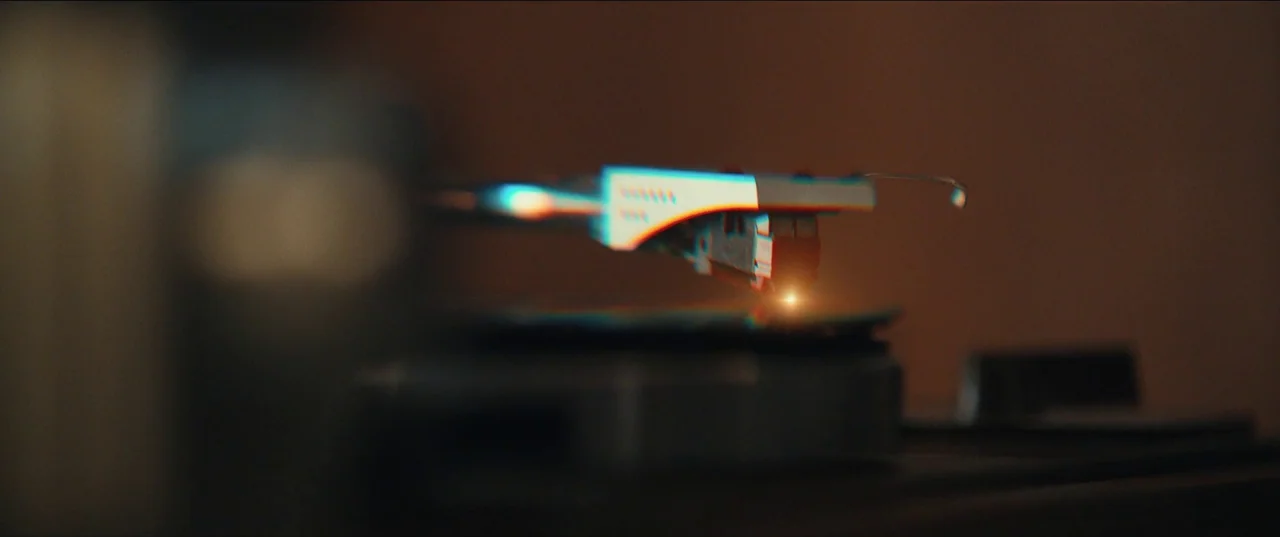 The Greatest Hits (2024) movie still: closeup — A dark indoor space contains a vintage turntable with a mechanical arm and needle. The stylus rests…