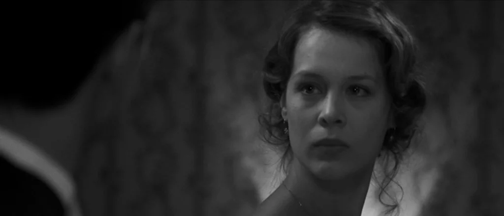 Frantz (2016) movie still: closeup — An interior space features patterned wallpaper in a dimly lit, high-contrast environment. A woman with curled…