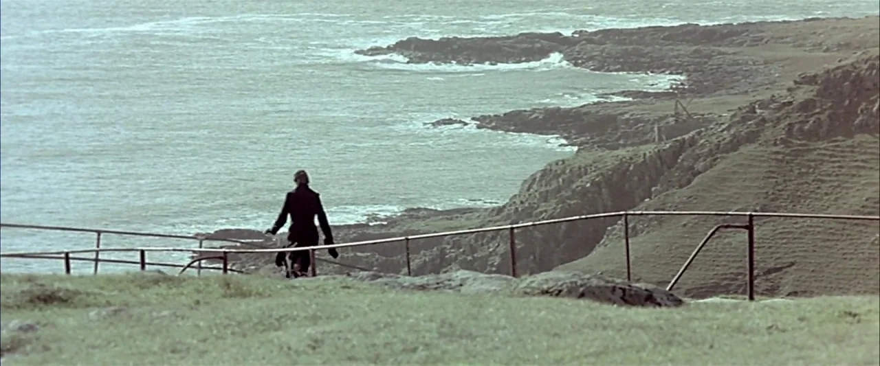 Breaking the Waves (1996) movie still: wide — A rugged coastline featuring grassy cliffs falling away toward a grey, choppy sea. A woman in a dark…
