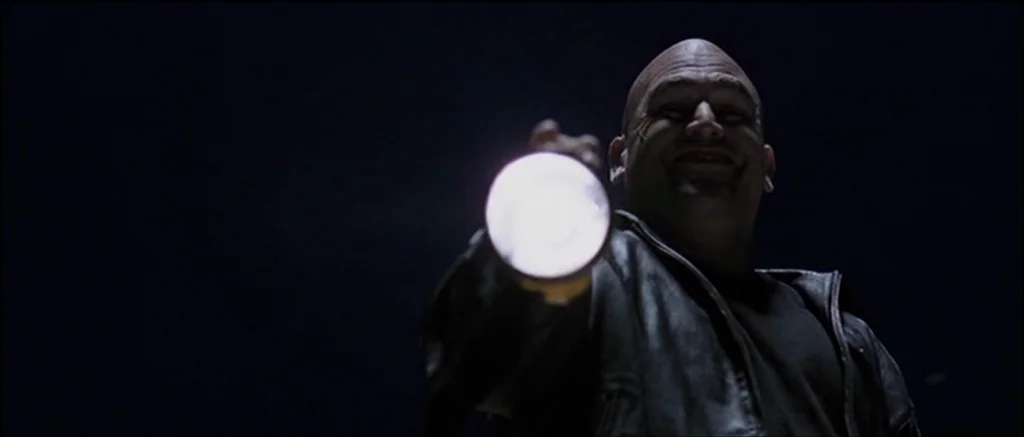 The Machinist (2004) movie still: medium — A dark environment surrounds a man in a black jacket. The man shines a bright flashlight directly at the…