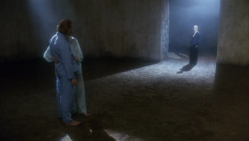 Edward II (1991) movie still: wide — A dark, cavernous space with rough, light-colored stone walls and a dirt floor. Two people stand embracing in…