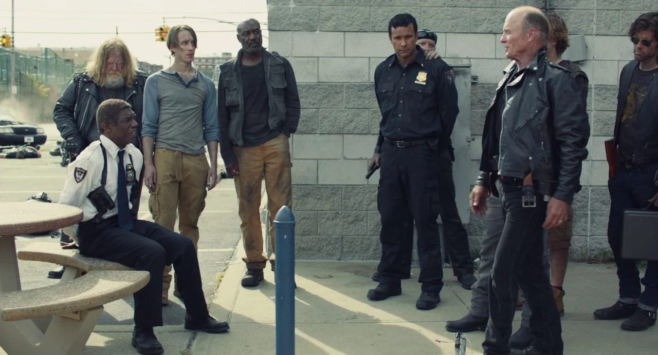 Cymbeline (2014) movie still: fullbody — A group of men stand on a grey concrete lot beside a masonry wall. An older man in a leather jacket stands…