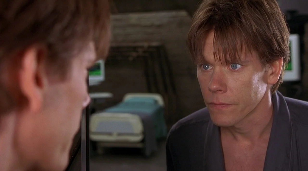Hollow Man (2000) movie still: medium — A hospital room interior features a blurred bed in the background and dark walls. A man stares directly at…