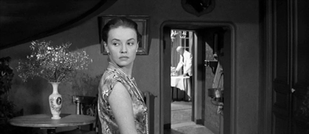 Les Amants (1958) movie still: medium — A dimly lit room features a woman in the foreground and a doorway. An elderly man stands in the distant room…