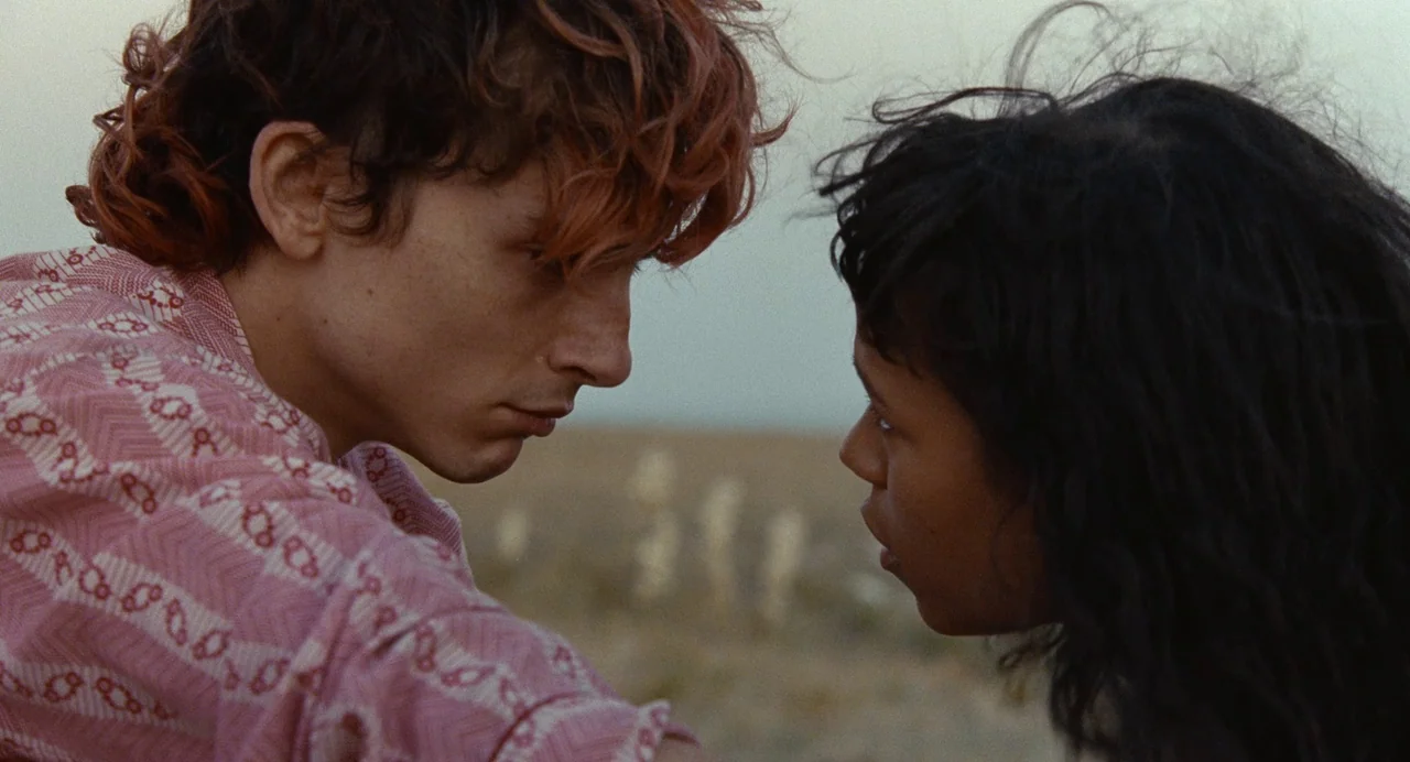 Bones and All (2022) movie still: closeup — A vast, arid landscape provides a muted backdrop for two individuals facing each other. The pair stands…