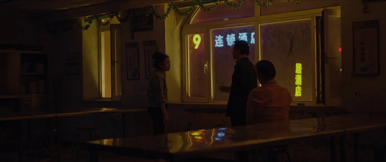 The Farewell (2019) movie still: medium — A dimly lit room features long metal tables and windows with visible neon signage outside. Three people…