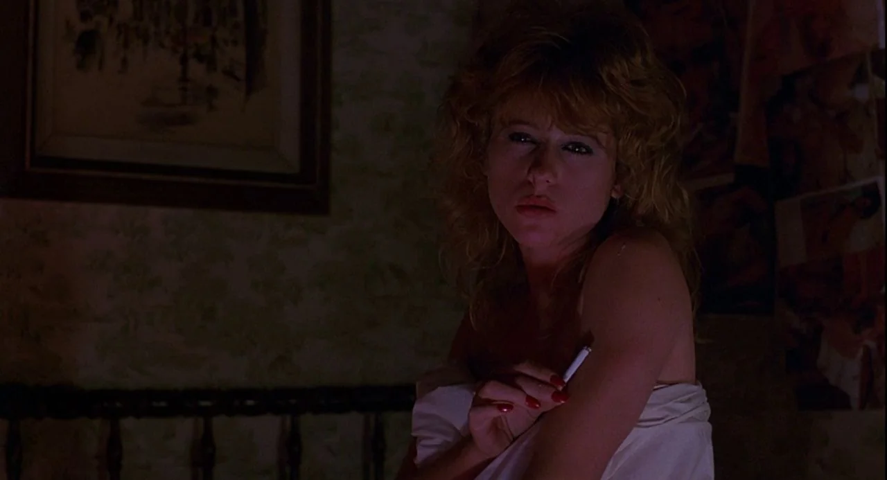 Psycho III (1986) movie still: medium — A dimly lit room with wallpapered walls and a framed picture hanging above. A woman sits on a bed covered by…