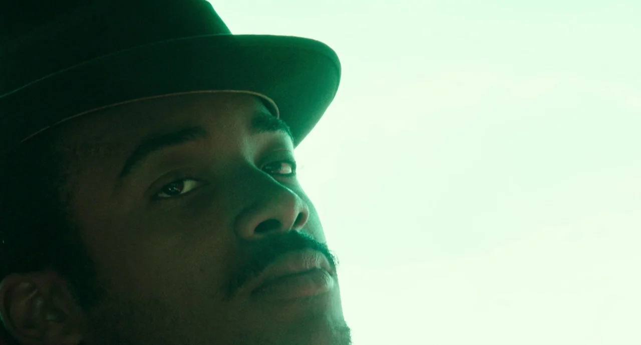 Detroit (2017) movie still: closeup — A close portrait of a man wearing a wide-brimmed hat against a bright background. The man looks toward the…