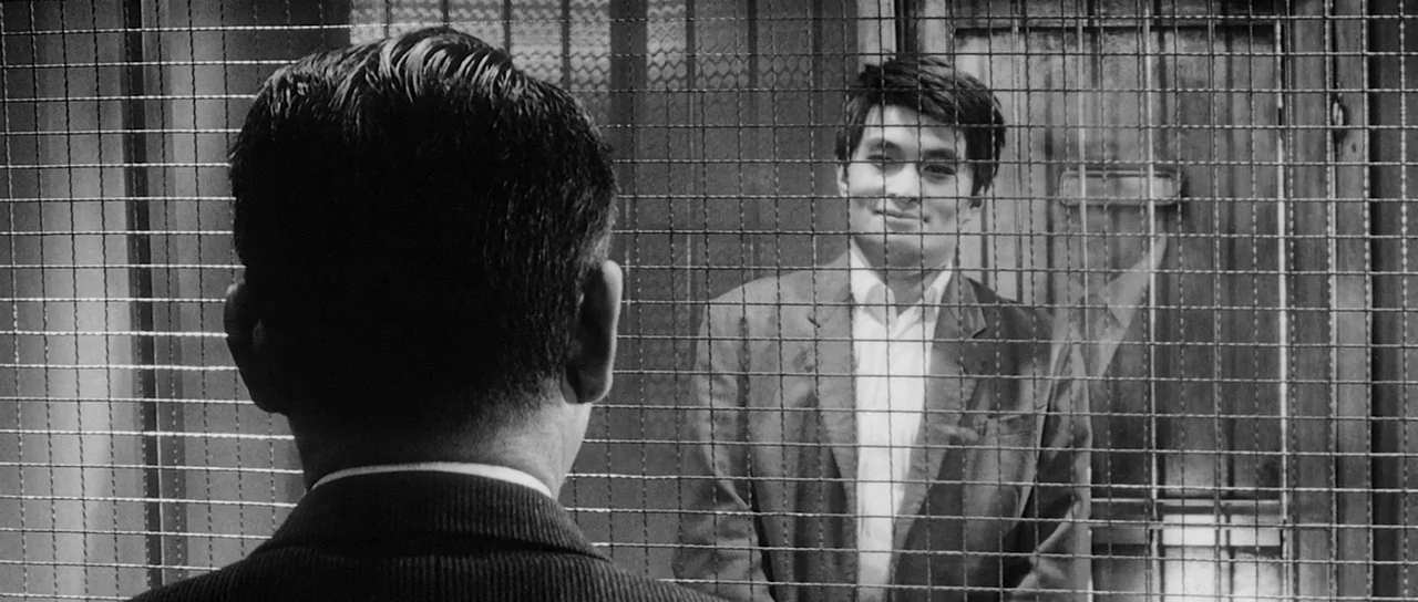 High and Low (1963) movie still: medium — A man stands behind a thick wire mesh screen in a sparse room. He wears a suit and smiles while facing an…