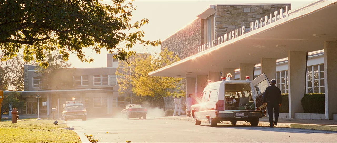 The Lovely Bones (2009) movie still: wide — The exterior of a 1970s stone hospital building at sunset with an ambulance parked. A man walks toward…