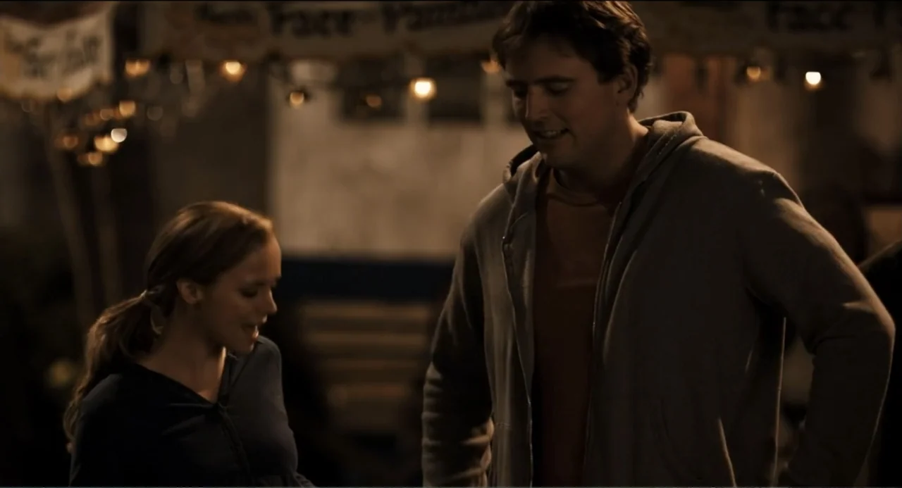 Todo es amor (2024) movie still: medium — An outdoor fairground setting at night featuring rows of string lights overhead. A man and a woman in…