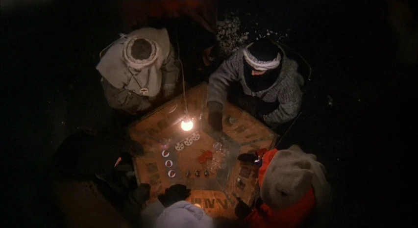 Quintet (1979) movie still: wide — Four people gather around a hexagonal wooden table inside a dimly lit dark space. They play a board game with…