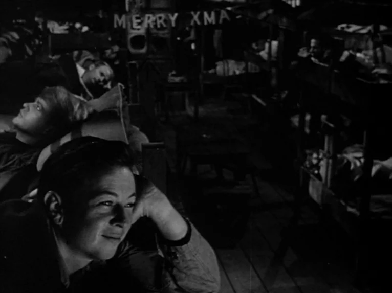 Stalag 17 (1953) movie still: medium — A crowded wooden barracks features multi-tiered bunk beds and a sign reading MERRY XMA. Two young men recline…