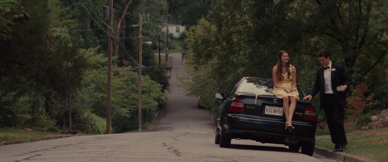 The Spectacular Now (2013) movie still: wide — A quiet residential street lined with mature trees features a car parked on the side. A young couple…