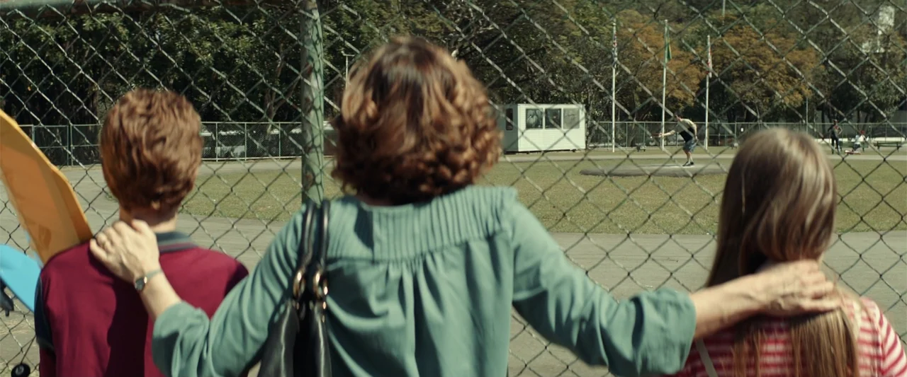 The Boy Who Killed My Parents (2021) movie still: medium — Three people stand together facing a wire fence overlooking an outdoor athletic field. A…