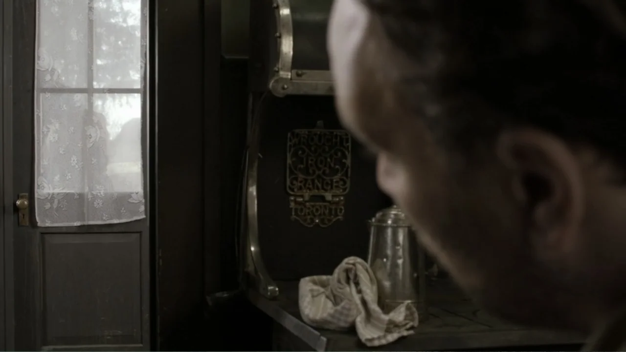 Dawn Rider (2012) movie still: closeup — A rustic kitchen interior features a cast iron stove and a lace-curtained door. A person in the foreground…