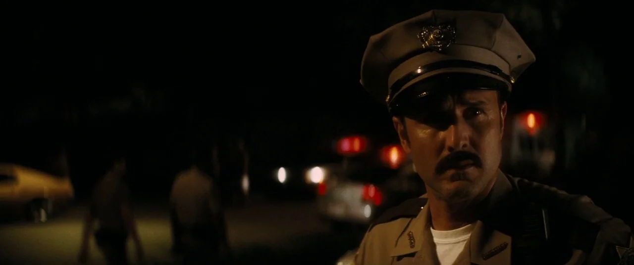 Scream 4 (2011) movie still: medium — A nighttime scene outdoors with out-of-focus vehicles and figures in the dark background. A police officer in…