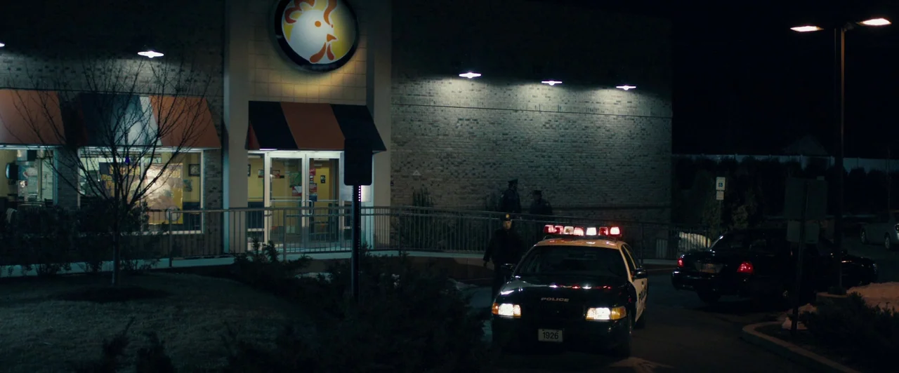 Compliance (2012) movie still: wide — A brick-walled fast food restaurant at night features orange awnings and a circular logo. A marked police…