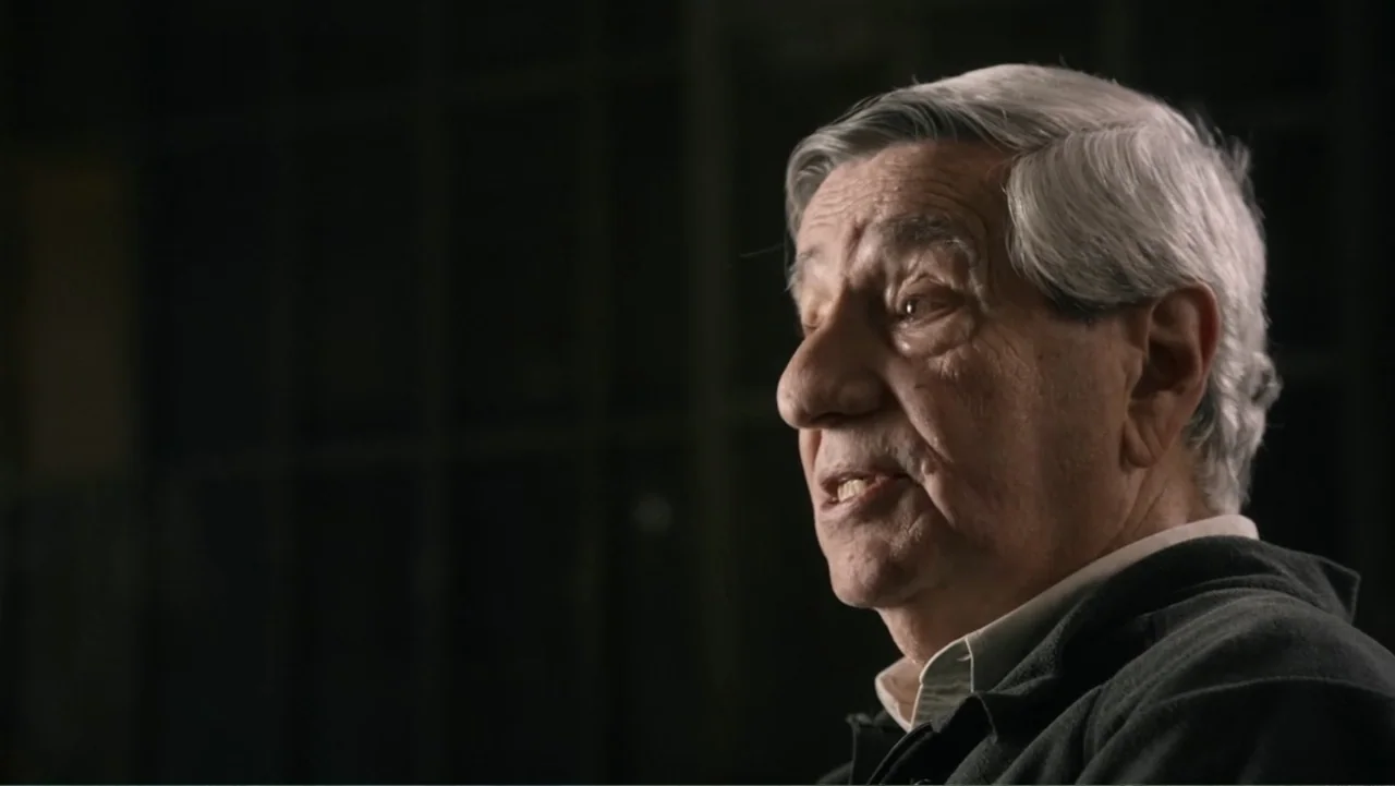 Transfers (2024) movie still: closeup — A dimly lit indoor space with an out-of-focus dark background behind the subject. An older man in a green…