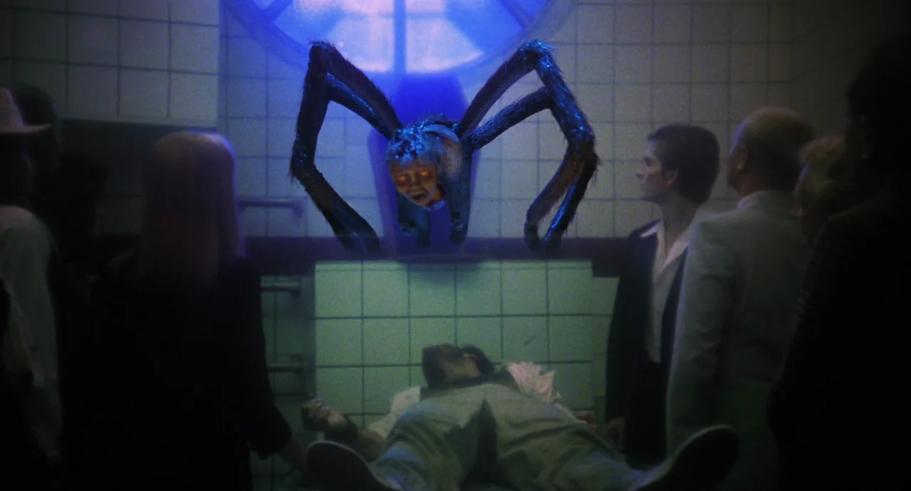 The Spider Labyrinth (1988) movie still: medium — A dark, tiled room features a low ceramic slab with a deceased body present. A creature with long…