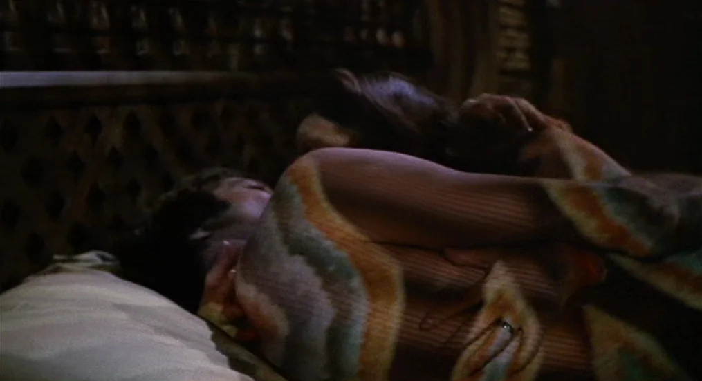 The Trip (1967) movie still: closeup — A dark, wood-paneled room interior features a bed with a detailed headboard. Two people embrace under a…