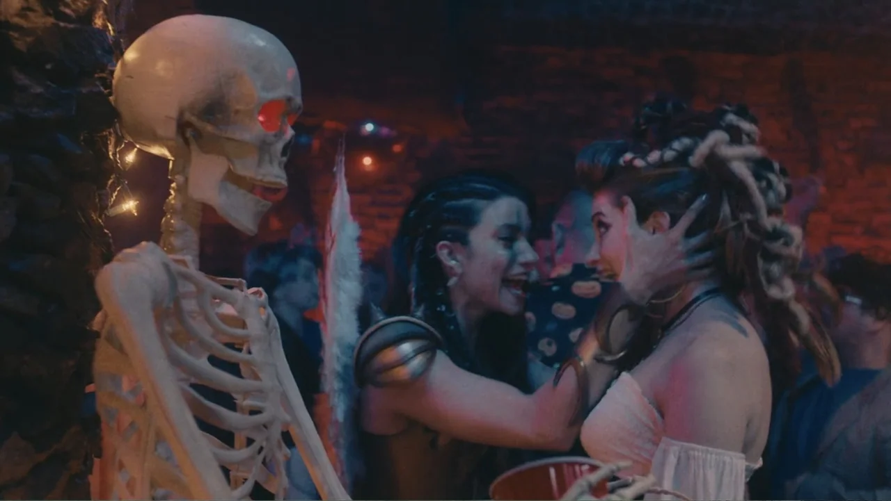 Terrifier 2 (2022) movie still: medium — A crowded dimly lit space with brick walls decorated with holiday string lights. Two women stand close…