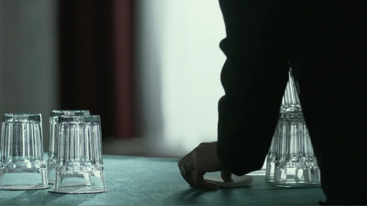 Confirmation (2016) movie still: medium — A dark table surface features several clear drinking glasses positioned in front of a window. A person…