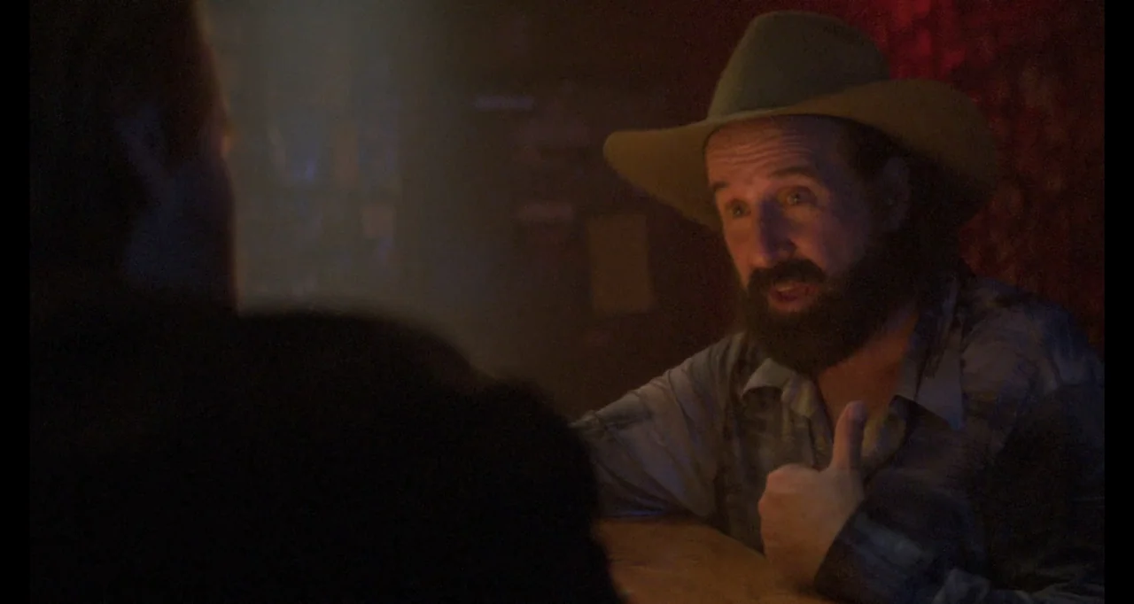 The Poison Rose (2019) movie still: medium — A dimly lit bar interior featuring dark wood surfaces and hazy ambient atmosphere. A man wearing a…