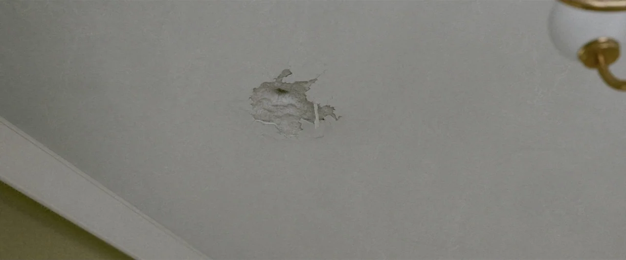 Voy a pasármelo bien (2022) movie still: closeup — A damaged white ceiling displays a jagged patch where the plaster has peeled away. The hole…