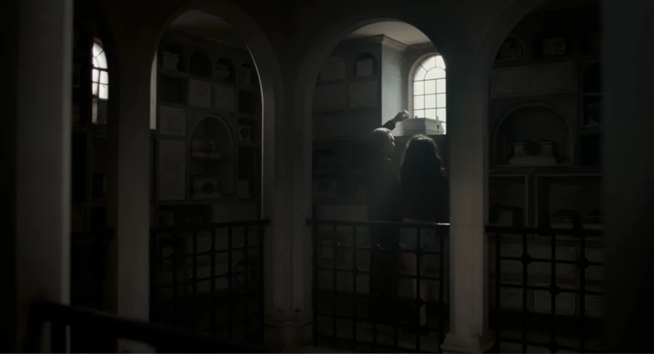 Back to Black (2024) movie still: medium — A stone mausoleum hallway features arched openings and rows of burial compartments. A man and a woman…